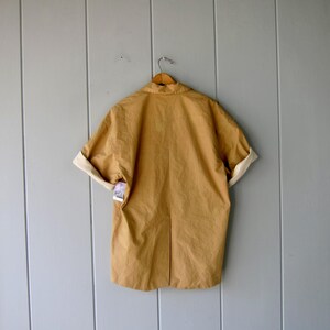 May include: A tan-colored short-sleeved jacket with rolled-up sleeves, revealing a cream-colored lining. The jacket is hanging on a wooden hanger against a light gray wooden wall. A tag is attached to the jacket.