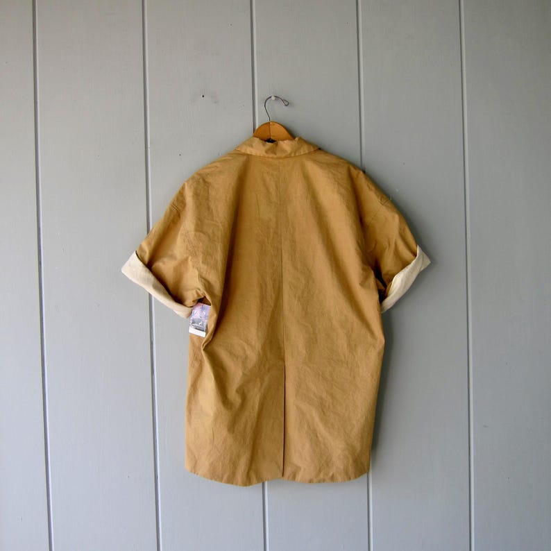 May include: A tan-colored short-sleeved jacket with rolled-up sleeves, revealing a cream-colored lining. The jacket is hanging on a wooden hanger against a light gray wooden wall. A tag is attached to the jacket.