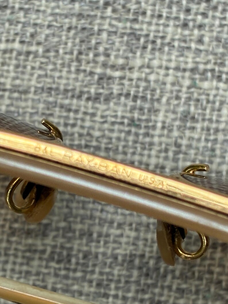 Puede incluir: Primer plano de unas gafas Ray-Ban USA. La montura es dorada con un patr&oacute;n marr&oacute;n claro y negro. El texto "RAY-BAN USA" es visible en la montura.