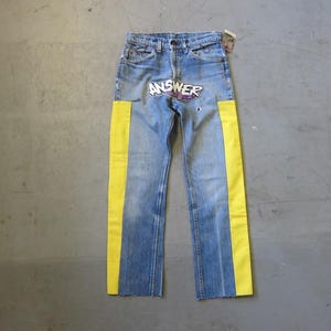 Pode incluir: Jeans azuis com faixas laterais amarelas brilhantes. Os jeans têm um visual desgastado com uma lavagem desbotada e um gráfico que diz "ANSWER" na frente. Os jeans têm um design clássico de cinco bolsos.