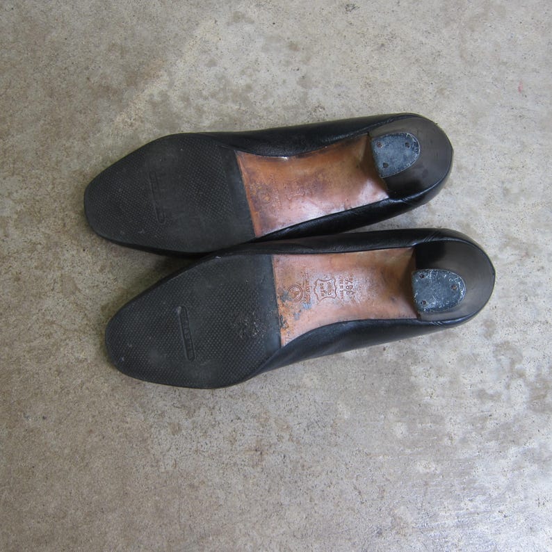 May include: A pair of black leather heeled shoes with a low heel. The shoes have a smooth leather upper and a rubber sole.