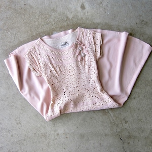 May include: A light pink top and skirt set. The top is made of a delicate, open-weave lace fabric with short sleeves and a round neckline. A small floral embellishment is on the right shoulder. The skirt is a solid, matching pink fabric.