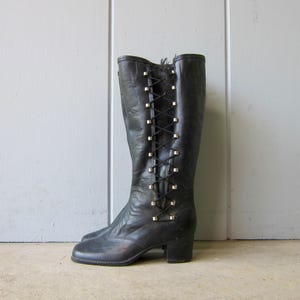 May include: A black knee-high boot featuring a side lace-up closure with thin black laces threaded through silver-toned metal eyelets. The boot has a rounded toe, a low block heel, and a smooth leather-like finish with visible natural creasing.