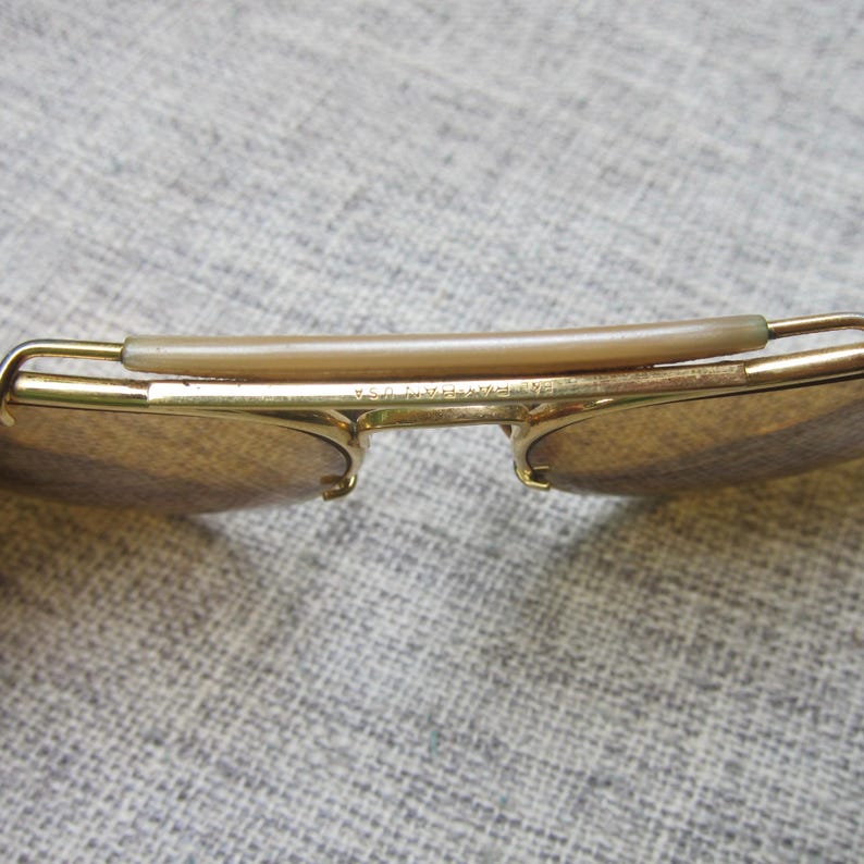 Puede incluir: Primer plano de gafas de sol doradas vintage con lentes marrones. Las gafas de sol tienen un estilo aviador cl&aacute;sico con puente y patillas. El texto "ERA PAT USA" es visible en la montura.