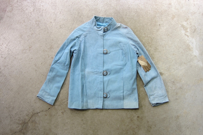 May include: A light blue jacket with a mandarin collar and four silver buttons. The jacket has long sleeves and a slightly fitted silhouette.