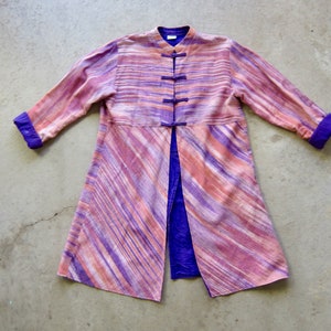 May include: A long, purple and pink striped coat with a mandarin collar and a front closure with buttons. The coat has a purple lining and is made of a soft, flowing fabric.