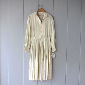 May include: A long-sleeved midi dress in a pale cream colour featuring vertical pinstripes in soft green and tan. The dress has a button-down front, a pointed collar, and a pleated skirt that falls from the waist, hanging on a wooden hanger.