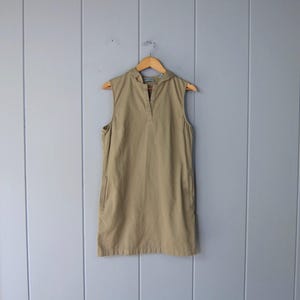 May include: A sleeveless, khaki-colored dress with a hood, hanging on a wooden hanger. The dress has a V-neck and two pockets. The background is a light gray wall with vertical wooden panels.