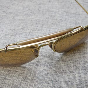 Puede incluir: Primer plano de gafas de sol doradas vintage con lentes marrones. Las gafas de sol tienen un estilo aviador cl&aacute;sico con puente y patillas. Las lentes son de color marr&oacute;n c&aacute;lido y la montura tiene un acabado brillante. El texto "USA" es visible.