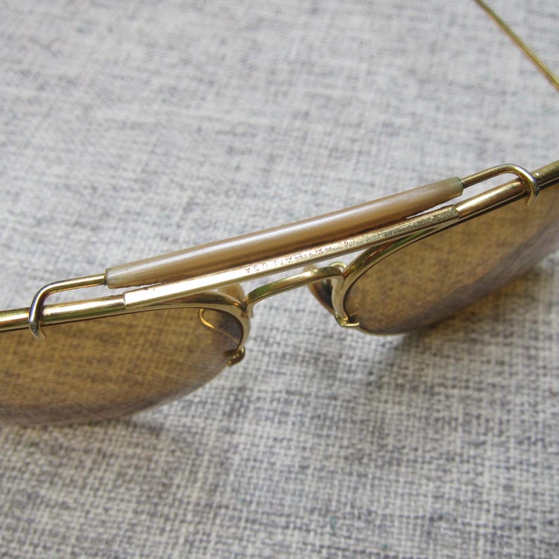 Puede incluir: Primer plano de gafas de sol doradas vintage con lentes marrones. Las gafas de sol tienen un estilo aviador cl&aacute;sico con puente y patillas. Las lentes son de color marr&oacute;n c&aacute;lido y la montura tiene un acabado brillante. El texto "USA" es visible.