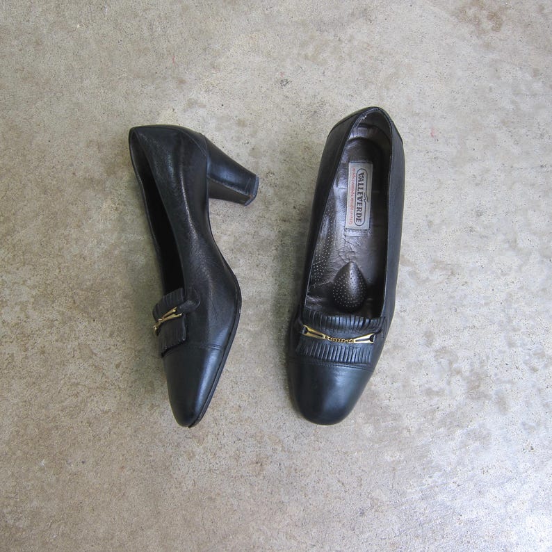 May include: A pair of black leather heeled shoes with a gold buckle detail. The shoes are labeled "Valleverde".
