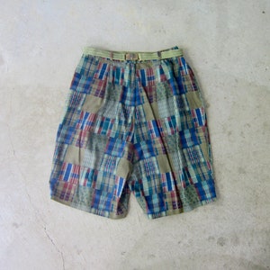 Può includere: Pantaloncini patchwork con cintura intrecciata. I pantaloncini presentano un mix di toppe a quadri e fantasia in blu, verde, rosso e beige. La cintura è in materiale intrecciato verde chiaro con fibbia rettangolare.