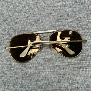 Puede incluir: Gafas de sol de aviador con montura dorada y lentes marrones. Las gafas tienen un doble puente y detalles blancos en las patillas. Las lentes reflejan el entorno.