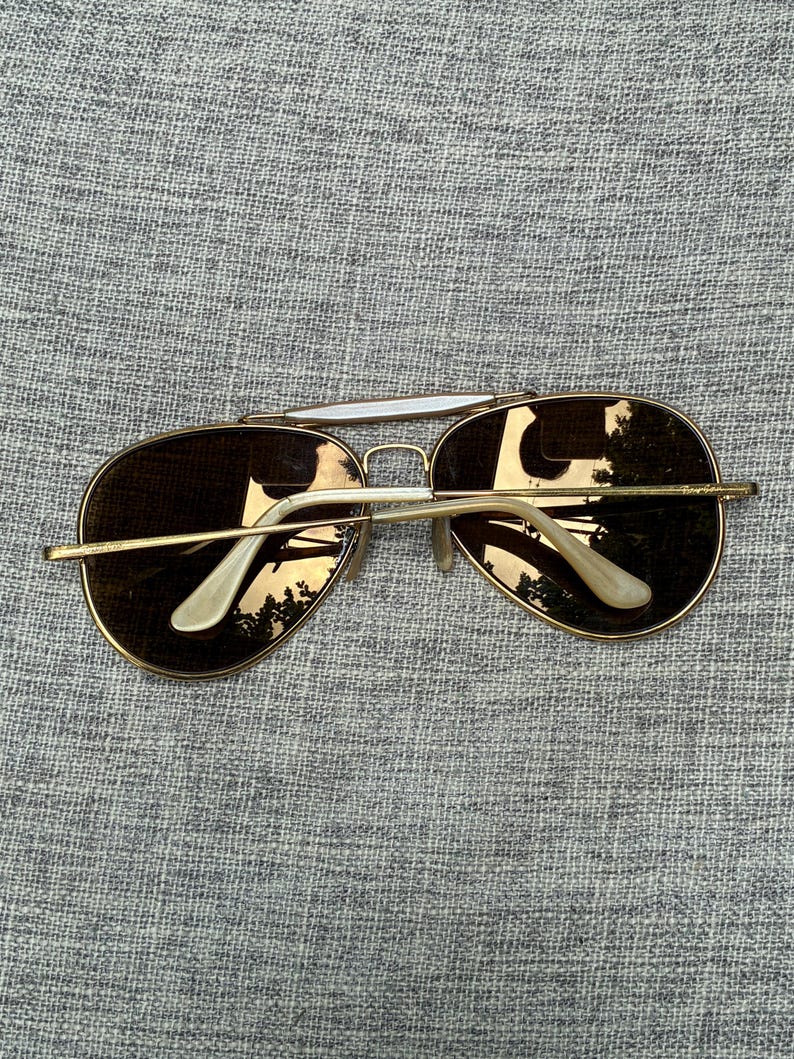 Puede incluir: Gafas de sol de aviador con montura dorada y lentes marrones. Las gafas tienen un doble puente y detalles blancos en las patillas. Las lentes reflejan el entorno.