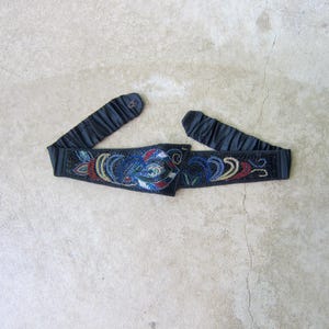 May include: A dark blue beaded belt with an elasticated band. The belt features a central panel with a colourful floral design in red, blue, and white beads. The elasticated ends are gathered.