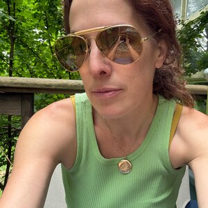 Puede incluir: Gafas de sol aviador doradas con lentes marrones. La persona lleva una camiseta sin mangas acanalada verde claro y un collar con colgante dorado con un dije circular. El fondo muestra follaje verde.