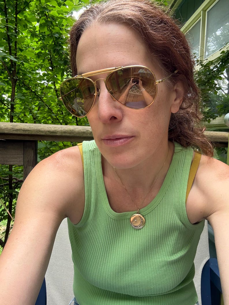 Puede incluir: Gafas de sol aviador doradas con lentes marrones. La persona lleva una camiseta sin mangas acanalada verde claro y un collar con colgante dorado con un dije circular. El fondo muestra follaje verde.