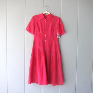 May include: A bright pink midi dress with short sleeves, a collared neckline, and a button-down front. The dress features a fitted bodice and a pleated skirt, hanging on a hanger against a light gray wall.