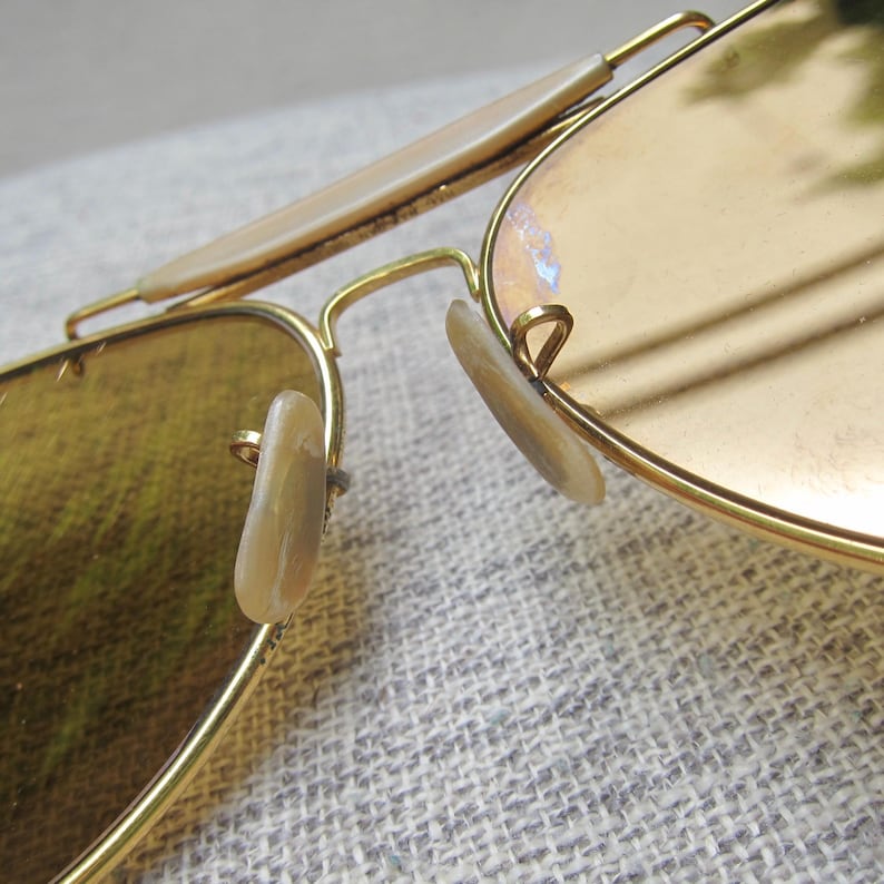 Puede incluir: Primer plano de gafas de sol de aviador con montura dorada y lentes marr&oacute;n claro. Las gafas de sol tienen un dise&ntilde;o cl&aacute;sico con doble puente y almohadillas nasales. Las lentes tienen un ligero tinte degradado.