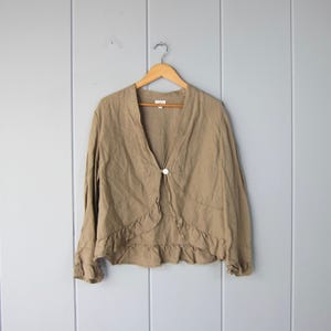May include: A tan linen jacket with long sleeves, a ruffled hem, and a single button fastening. The jacket is suspended on a wooden hanger against a pale blue wall.