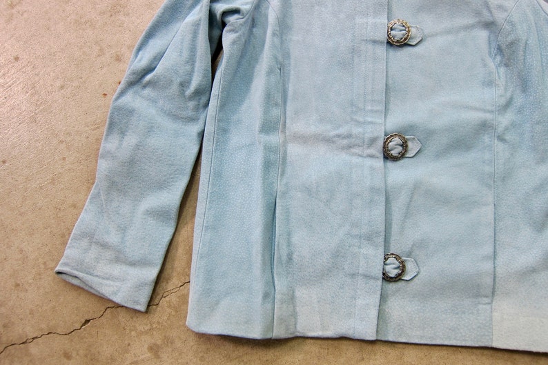May include: A light blue jacket with long sleeves and three decorative buttons.