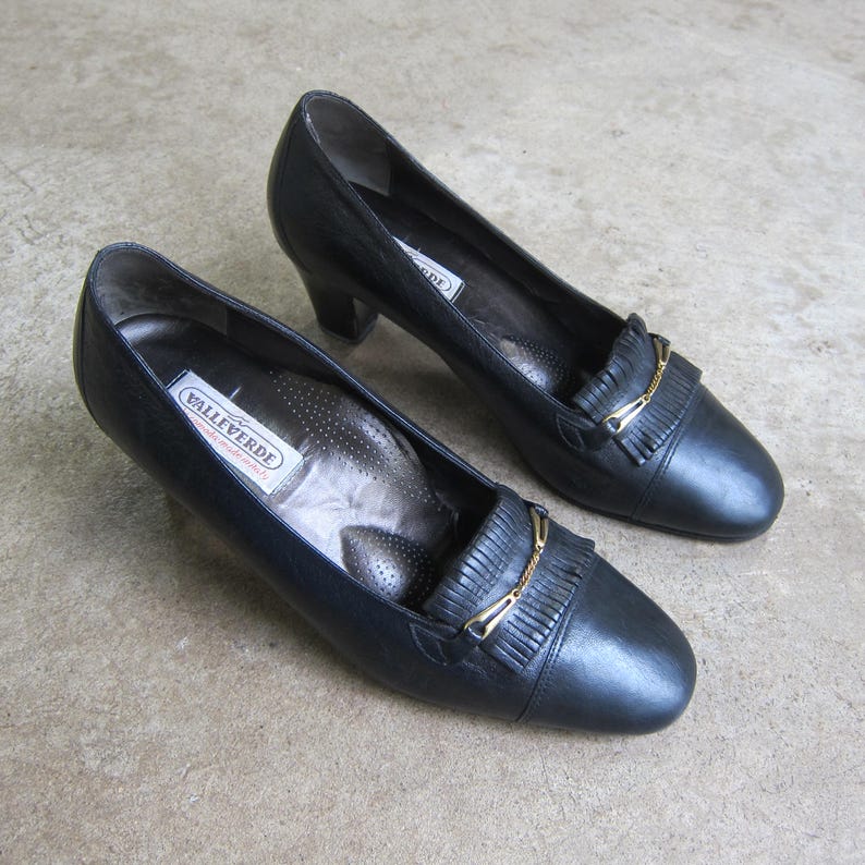 May include: A pair of black leather heeled shoes with gold accents and a fringe detail. The shoes are labeled "Valleverde" on the insole.