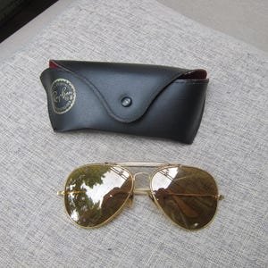 Puede incluir: Gafas de sol aviador con montura dorada y lentes marrones, junto a un estuche de cuero negro. El estuche tiene un cierre de bot&oacute;n a presi&oacute;n y un interior rojo. Las gafas est&aacute;n sobre una superficie gris.