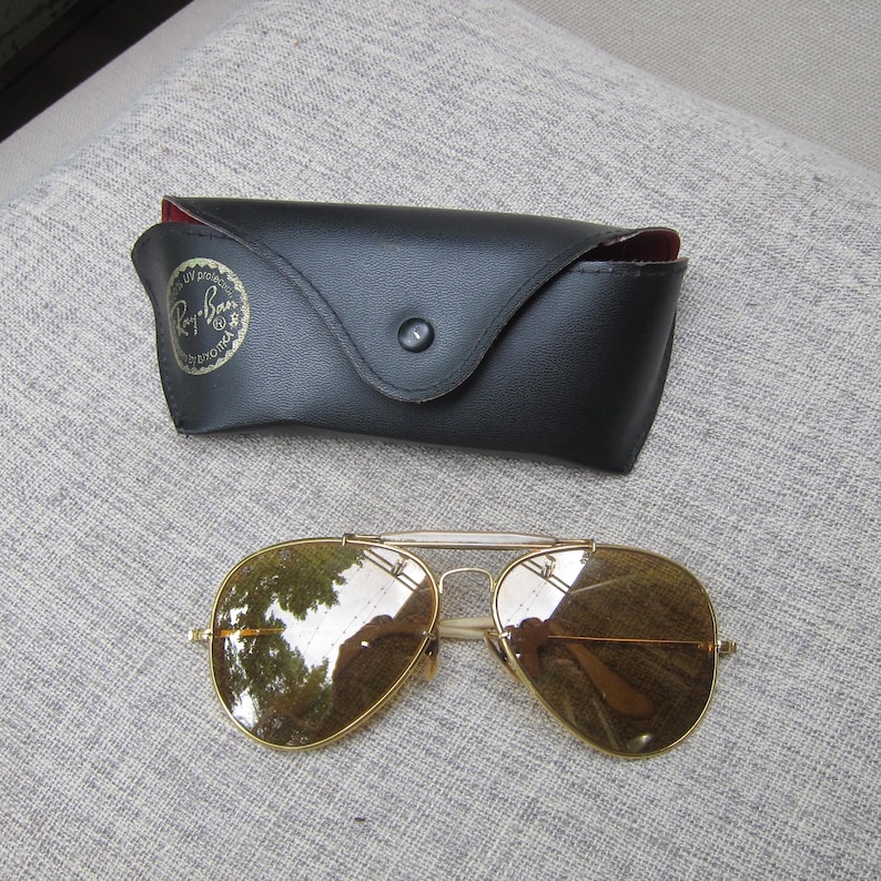 Puede incluir: Gafas de sol aviador con montura dorada y lentes marrones, junto a un estuche de cuero negro. El estuche tiene un cierre de bot&oacute;n a presi&oacute;n y un interior rojo. Las gafas est&aacute;n sobre una superficie gris.