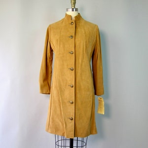 May include: A long-sleeved, tan suede coat with a mandarin collar and button closure. The coat has a straight silhouette and is displayed on a mannequin.
