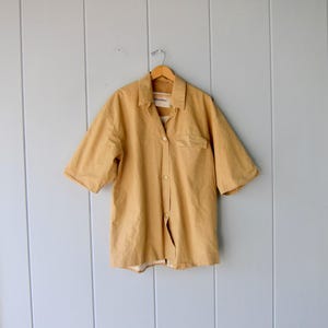May include: A short-sleeved, button-down shirt in a mustard yellow color. The shirt has a chest pocket on the left side and is hanging on a wooden hanger. The background is a light blue wall with vertical wooden panels.