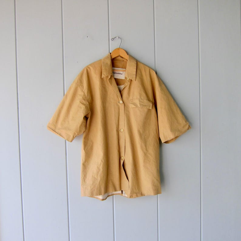 May include: A short-sleeved, button-down shirt in a mustard yellow color. The shirt has a chest pocket on the left side and is hanging on a wooden hanger. The background is a light blue wall with vertical wooden panels.