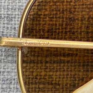 Puede incluir: Primer plano de un broche dorado con un dise&ntilde;o circular. El broche presenta un fondo marr&oacute;n tejido y un marco dorado. El texto "Roger Jean" est&aacute; inscrito en la barra dorada.