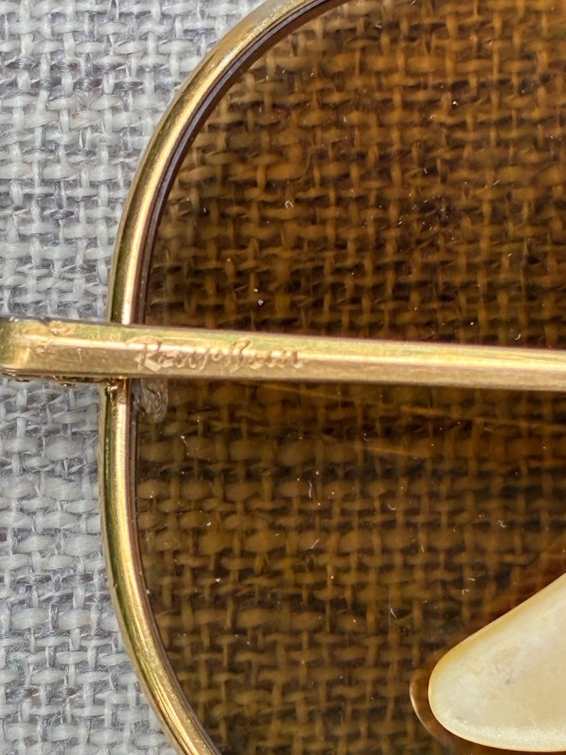 Puede incluir: Primer plano de un broche dorado con un dise&ntilde;o circular. El broche presenta un fondo marr&oacute;n tejido y un marco dorado. El texto "Roger Jean" est&aacute; inscrito en la barra dorada.