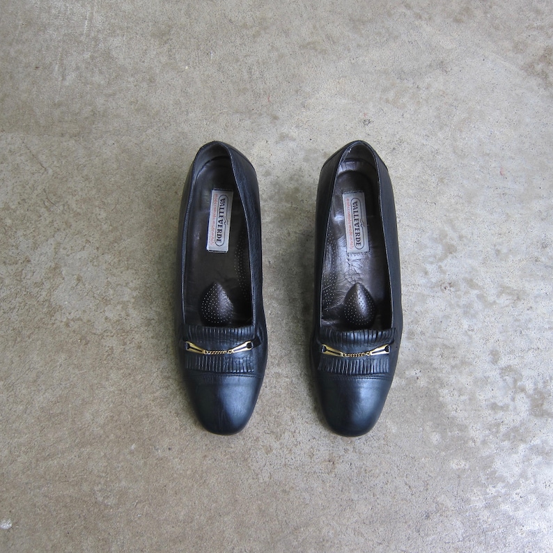 May include: A pair of black leather heeled shoes with gold accents. The shoes have a pointed toe and a small gold buckle on the front. The brand name "VILLATTIVA" is printed on the inside of each shoe.