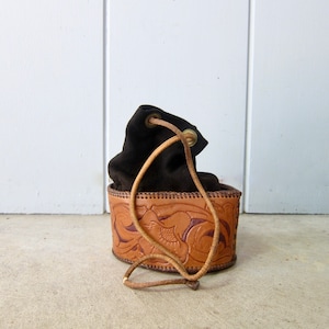 Könnte beinhalten: Eine braune Ledertasche mit Kordelzug und einem geprägten Design. Die Tasche hat einen braunen Lederriemen und eine Messing-Schnalle.