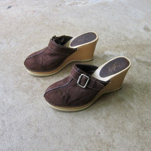May include: A pair of brown suede platform clogs with a wooden wedge heel. The clogs have a buckle strap and the brand name "Candice" is printed on the insole.