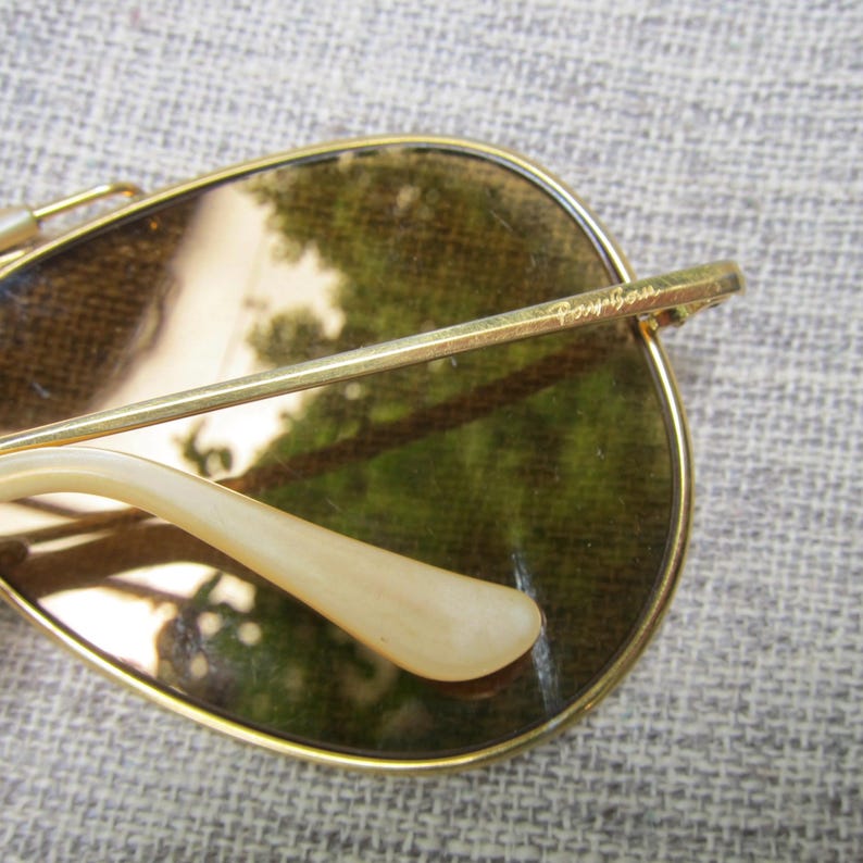 Puede incluir: Primer plano de gafas de sol con montura dorada y lentes marrones. El logotipo de Ray-Ban es visible en la patilla. Las patillas son de color crema claro. Las lentes reflejan una escena borrosa al aire libre.