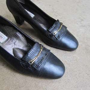 May include: A pair of black leather heeled shoes with a gold chain and fringe detail. The shoes have a pointed toe and a low heel.