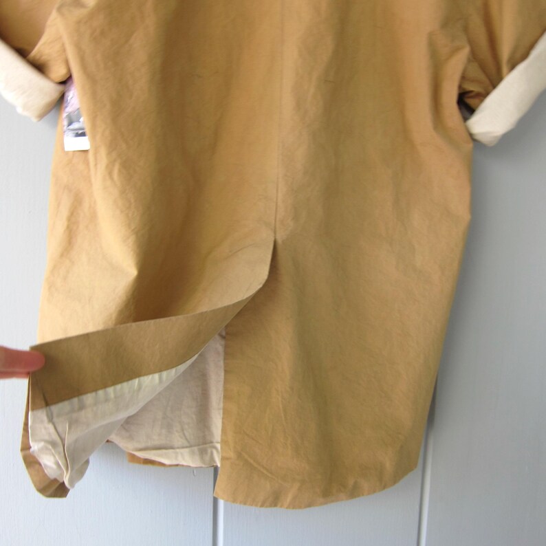 May include: Close-up of a tan-colored jacket with rolled-up sleeves. The jacket has a split in the back, revealing a lighter-colored lining. The cuffs are a cream color. A small tag is visible near the sleeve.