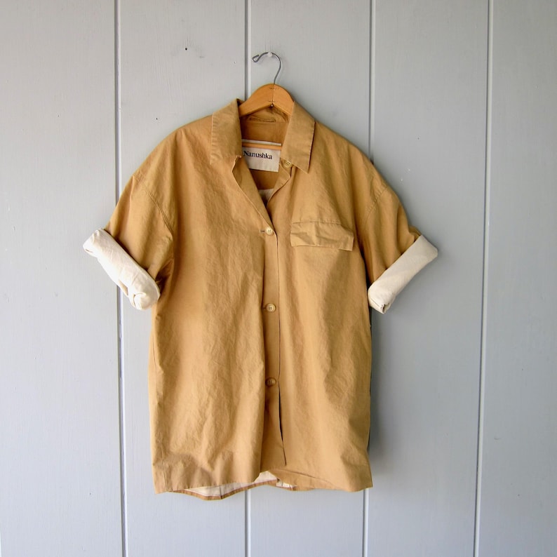 May include: A tan button-down shirt with rolled-up sleeves, hanging on a wooden hanger. The sleeves are cuffed to reveal a cream-colored lining. The shirt has a chest pocket and the brand name "Nanushka" is visible on the collar.
