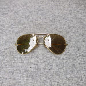 Puede incluir: Gafas de sol de aviador con montura dorada y lentes marrones. Las gafas tienen un doble puente y una forma cl&aacute;sica de l&aacute;grima. Las lentes reflejan el entorno. La montura es de color dorado brillante.