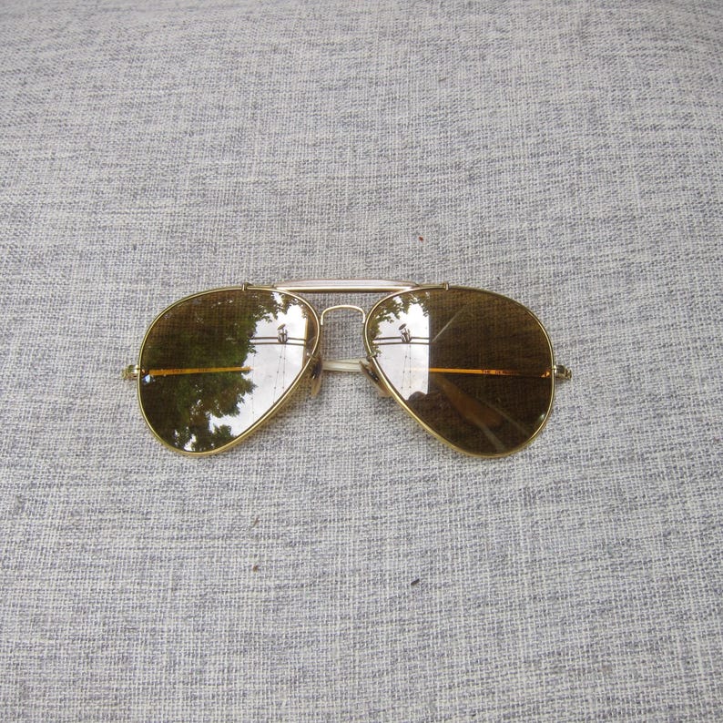 Puede incluir: Gafas de sol de aviador con montura dorada y lentes marrones. Las gafas tienen un doble puente y una forma cl&aacute;sica de l&aacute;grima. Las lentes reflejan el entorno. La montura es de color dorado brillante.