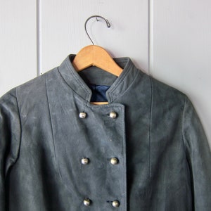Peut inclure: Une veste en daim gris avec un col mandarin et une fermeture &agrave; double boutonnage. La veste a des boutons argent&eacute;s et est suspendue &agrave; un cintre en bois.