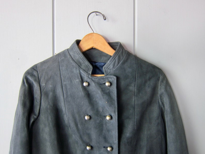 Peut inclure: Une veste en daim gris avec un col mandarin et une fermeture &agrave; double boutonnage. La veste a des boutons argent&eacute;s et est suspendue &agrave; un cintre en bois.