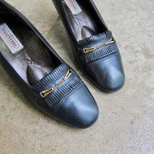 May include: A pair of black leather heeled shoes with gold chain accents and fringe detailing. The shoes have a pointed toe and a low heel. The brand name "Valleverde" is visible on the insole.