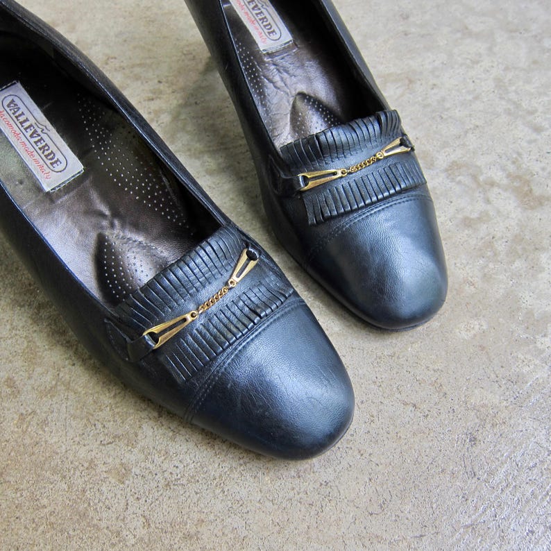 May include: A pair of black leather heeled shoes with gold chain accents and fringe detailing. The shoes have a pointed toe and a low heel. The brand name "Valleverde" is visible on the insole.