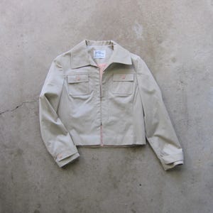 Puede incluir: Una chaqueta beige con cremallera, dos bolsillos de solapa y una etiqueta que dice "Kline Boutique".