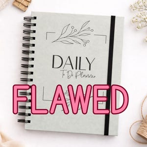 May include: A light grey spiral-bound planner with the words "DAILY To Do Planner" and a floral design. A black elastic band holds the planner closed. The word "FLAWED" is written across the front in pink letters.