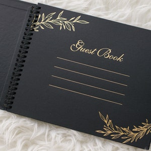 May include: Black guest book with gold foil lettering and floral designs. The book has a spiral binding and is open to a blank page with lines for writing.