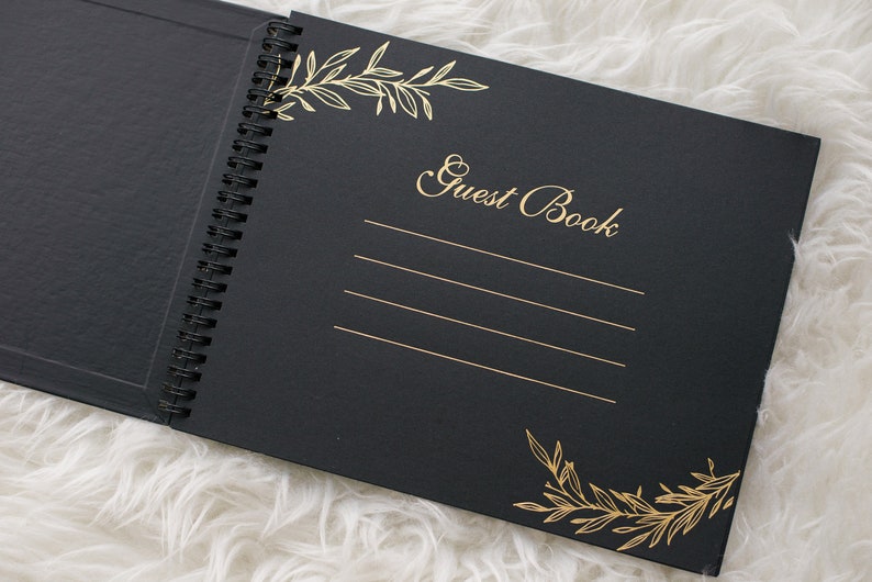 May include: Black guest book with gold foil lettering and floral designs. The book has a spiral binding and is open to a blank page with lines for writing.